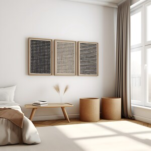 May include: Three framed abstract art prints with black and beige geometric patterns hang above a wooden bench. Two woven cylindrical baskets sit on the floor. The room has a neutral color palette with a beige rug and a white wall.