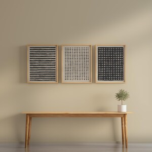 May include: Three framed abstract art prints with black and white geometric patterns hang above a wooden bench. The prints feature horizontal lines, a grid, and a checkered design. A small potted plant sits on the bench.