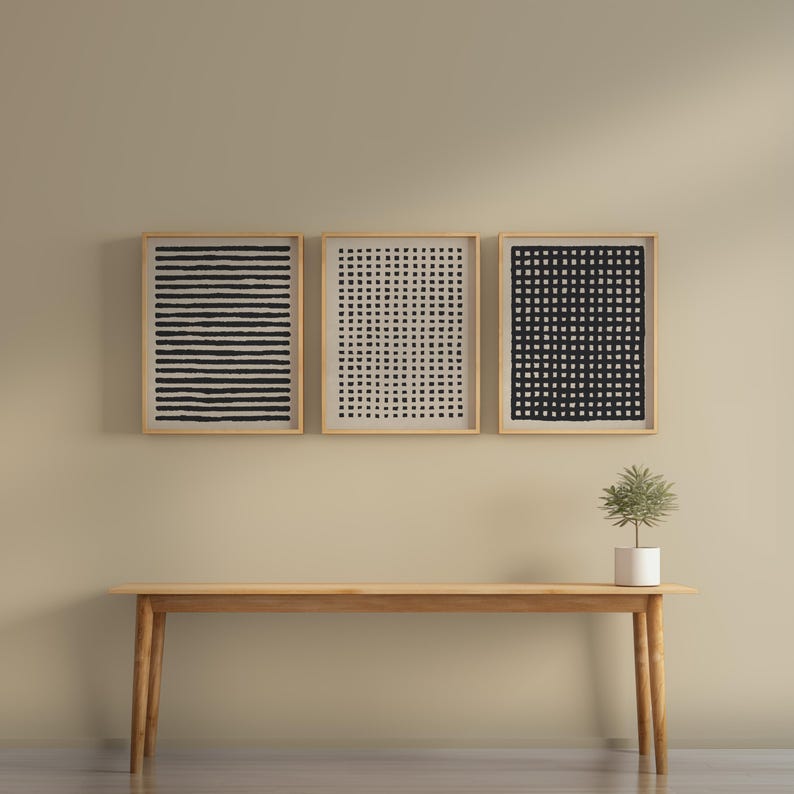 May include: Three framed abstract art prints with black and white geometric patterns hang above a wooden bench. The prints feature horizontal lines, a grid, and a checkered design. A small potted plant sits on the bench.