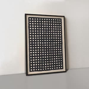 May include: Black and white framed artwork. The artwork features a grid pattern of black squares on a cream background. The frame is black and the artwork is leaning against a white wall.