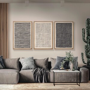 May include: Three framed abstract art prints with black and white geometric patterns hang above a gray sectional sofa. The prints feature horizontal lines, dots, and a grid design. A modern living room setting.