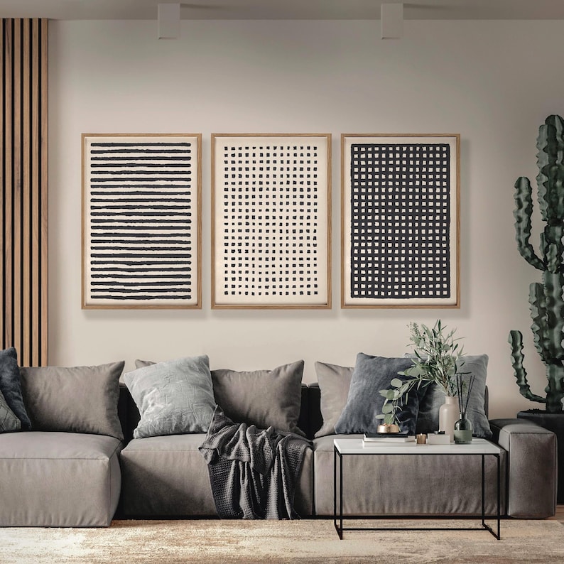 May include: Three framed abstract art prints with black and white geometric patterns hang above a gray sectional sofa. The prints feature horizontal lines, dots, and a grid design. A modern living room setting.