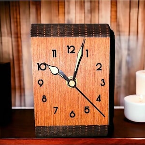 May include: A wooden clock with black hands and Roman numeral markings. The clock is made of dark wood with a light wood face. The clock is sitting on a wooden surface.
