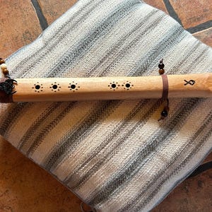 May include: A handcrafted wooden flute with a natural finish, featuring decorative holes and a leather strap. The flute rests on a striped pillow. The instrument has a small wooden block at the end and is adorned with beads.