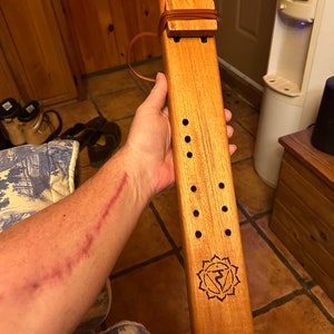 May include: A wooden flute with a brown leather strap and a carved symbol of a chakra on the bottom. The flute has several holes along the side.