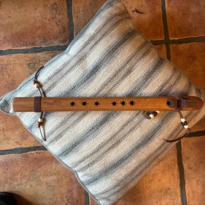 May include: A wooden flute with a sunburst design is displayed on a striped pillow. The flute is brown with six star-shaped cutouts and leather accents. The pillow has a neutral color palette.