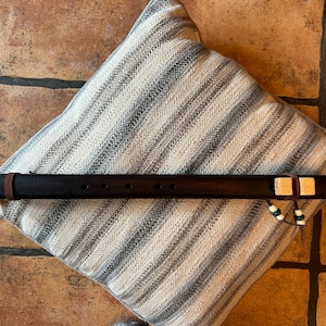 May include: A dark brown wooden flute with six finger holes, resting on a striped pillow. The flute has a leather strap and decorative beads. The pillow has a neutral color scheme with gray and white stripes.