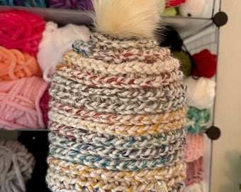 Może przedstawiać: Ręcznie robiona czapka typu beanie z kremowym pomponem z imitacji futra. Czapka ma teksturowany, wielokolorowy wzór z odcieniami kremu, czerwieni, żółci i turkusu. Do brzegu przymocowana jest mała metka. Czapka jest prezentowana na tle kolorowej przędzy.