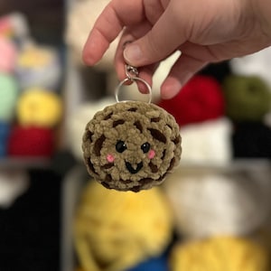 May include: A brown crocheted keychain shaped like a cookie with a smiling face and pink cheeks.