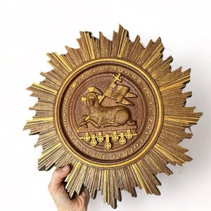 Op de afbeelding: Een houten, cirkelvormig religieus embleem met een gedetailleerde reliëfsnede. Het embleem toont een lam, een spandoek en tekst binnen een zonnestraalframe. Het kleurenpalet omvat tinten bruin en goud.