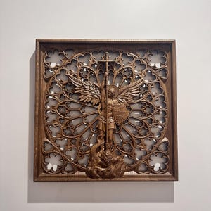Op de afbeelding: Houten wandkunst met een gedetailleerd reliëf van de aartsengel Michaël. Het kunstwerk is geplaatst in een vierkante lijst en heeft een decoratieve, opengewerkte achtergrond. De aartsengel wordt afgebeeld met vleugels, een schild en een speer, vechtend tegen een slang.