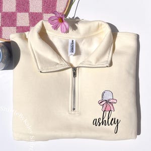 May include: Cream-colored quarter-zip sweatshirt with a silver zipper. The sweatshirt features an embroidered design of a baby bonnet with a pink bow and the name "ashley" in cursive. A pink flower rests on the folded collar.