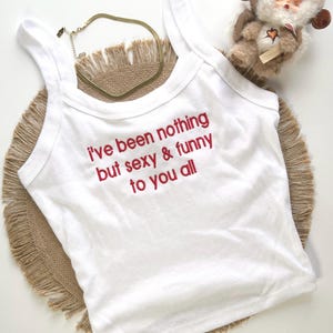 May include: White tank top with the embroidered text "i've been nothing but sexy & funny to you all" in red. A gold-colored necklace and a small doll with a furry hood are also visible.