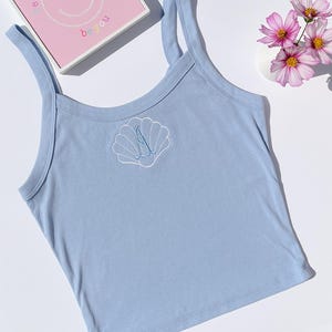 Puede incluir: Una camiseta sin mangas azul claro con un diseño de concha bordada en blanco. La camiseta tiene tirantes finos y un escote redondo. Una caja rosa con una carita sonriente y las palabras "be you" es visible en el fondo. También hay flores rosas.