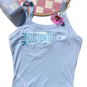 May include: Light blue tank top with a pennant design that reads "CUSTOM TEXT". The pennant is decorated with a floral pattern and a star. A pink flower and a blue mug with white bows are also in the image.