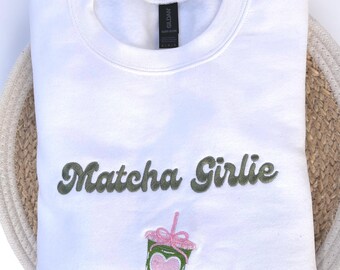 May include: White sweatshirt with the embroidered text "Matcha Girlie" in olive green. Below the text is an embroidered green matcha drink with a pink heart and straw. The sweatshirt is folded on a woven basket.