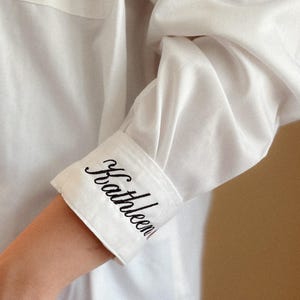 Puede incluir: Primer plano de una manga de camisa blanca con el nombre "Kathleen" bordado en escritura negra en el puño. El puño está doblado, revelando el nombre bordado. La camisa es de tela blanca lisa.