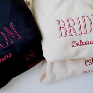 May include: A black hoodie with pink embroidered text reading GROOM, Deloera, and est 2023, alongside a cream hoodie with pink embroidered text reading BRIDE, Deloera, and est 2023. Both garments feature long sleeves and hoods with drawstrings.