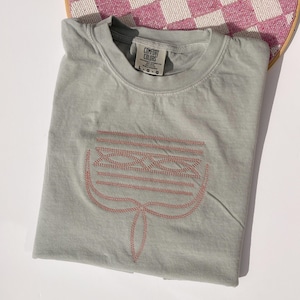 Puede incluir: Camiseta verde salvia con cuello redondo y mangas cortas. Presenta un diseño bordado en rosa rojizo, una forma floral o abstracta estilizada. La camiseta está doblada cuidadosamente.