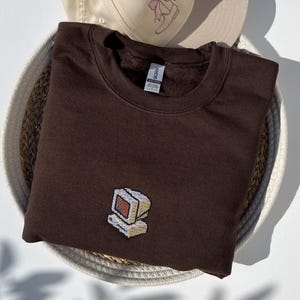 May include: A brown sweatshirt with a small embroidered computer design. The sweatshirt is folded in a woven basket, with a beige baseball cap with a pink bow design in the background.