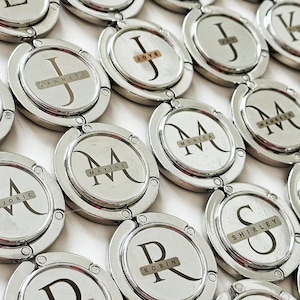 May include: A collection of silver-toned purse hangers, each featuring a circular design with a name or initial. The names are printed on small rectangular labels. The hangers are arranged on a white surface.