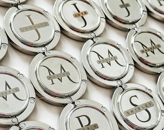 May include: A collection of silver-toned purse hangers, each featuring a circular design with a name or initial. The names are printed on small rectangular labels. The hangers are arranged on a white surface.