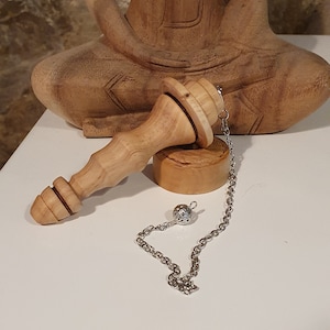 Op de afbeelding: Een houten slinger met een zilveren ketting en een kleine zilveren bal aan het uiteinde. De slinger rust op een houten voet. De slinger bevindt zich voor een houten beeld van een zittende Boeddha.