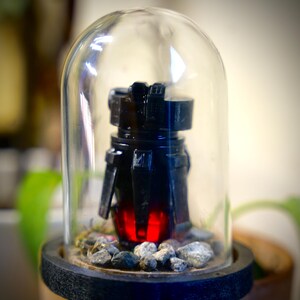 May include: A black, futuristic-looking object with a red glowing core, encased in a clear glass dome. The object sits on a bed of small gray stones, mounted on a dark wooden base. The dome is clear, allowing a full view of the object.