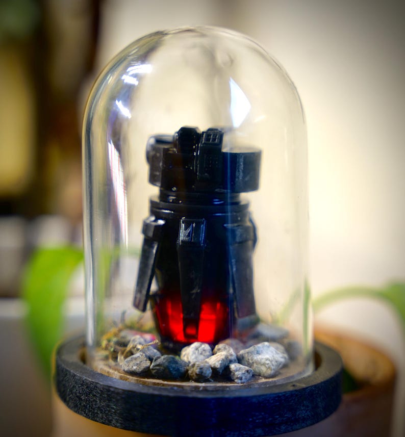 May include: A black, futuristic-looking object with a red glowing core, encased in a clear glass dome. The object sits on a bed of small gray stones, mounted on a dark wooden base. The dome is clear, allowing a full view of the object.