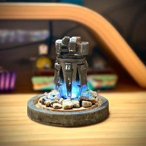 May include: A detailed, futuristic-looking sculpture with a dark gray, metallic finish. The sculpture has a cylindrical body with angular features and is illuminated by a bright blue light emanating from the base. The base is a circular platform with small rocks.