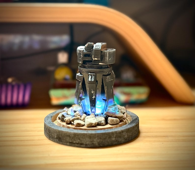 May include: A detailed, futuristic-looking sculpture with a dark gray, metallic finish. The sculpture has a cylindrical body with angular features and is illuminated by a bright blue light emanating from the base. The base is a circular platform with small rocks.