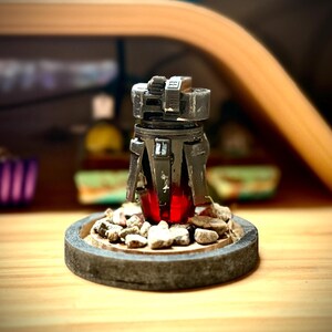 May include: A detailed, dark gray miniature sculpture with a red glowing core, set on a stone-like base with small rocks. The sculpture has a mechanical design, with visible details and textures. The base is approximately 3 inches in diameter.