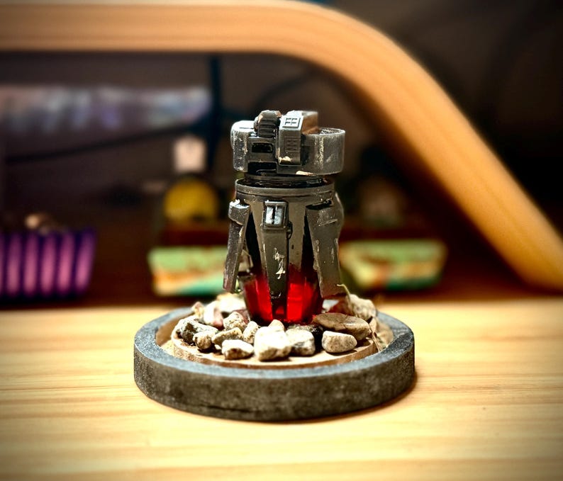 May include: A detailed, dark gray miniature sculpture with a red glowing core, set on a stone-like base with small rocks. The sculpture has a mechanical design, with visible details and textures. The base is approximately 3 inches in diameter.