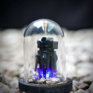 May include: A black robot figurine inside a clear glass dome. The robot has a boxy design with visible details. The base of the dome is a dark gray color, and the robot is illuminated with blue light.