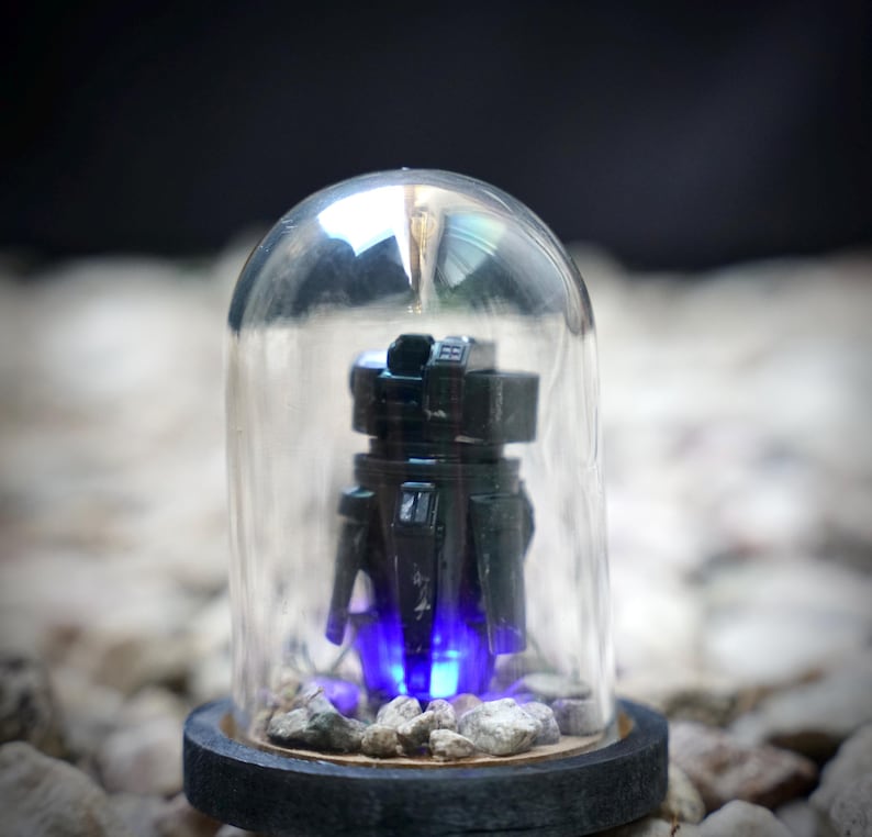 May include: A black robot figurine inside a clear glass dome. The robot has a boxy design with visible details. The base of the dome is a dark gray color, and the robot is illuminated with blue light.