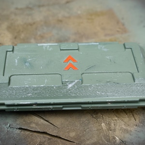 May include: A rectangular, olive-green container with a hinged lid. The lid features two orange upward-pointing arrows. The container has a textured surface and appears to be made of plastic or a similar material. The container is resting on a weathered surface.
