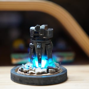 May include: A futuristic-looking, dark gray sculpture with a cylindrical body and angular details. The sculpture emits a bright blue light from within, resting on a circular base with small rocks. The overall design suggests a sci-fi or fantasy theme.
