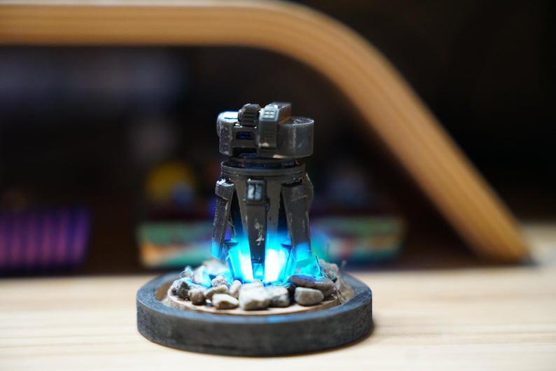 May include: A futuristic-looking, dark gray sculpture with a cylindrical body and angular details. The sculpture emits a bright blue light from within, resting on a circular base with small rocks. The overall design suggests a sci-fi or fantasy theme.