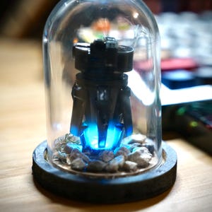 May include: A black, futuristic-looking sculpture with a bright blue light emanating from its base, encased in a clear glass dome. The sculpture sits on a wooden base with small rocks around it. The overall aesthetic is modern and artistic.