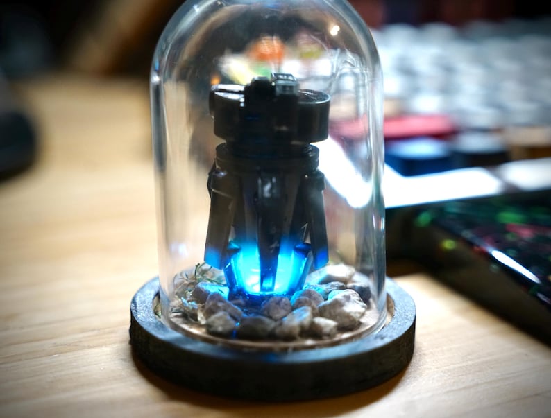 May include: A black, futuristic-looking sculpture with a bright blue light emanating from its base, encased in a clear glass dome. The sculpture sits on a wooden base with small rocks around it. The overall aesthetic is modern and artistic.