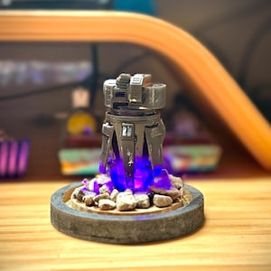 May include: A detailed, gray and black miniature sculpture with a futuristic design. The sculpture sits on a circular base with small rocks and emits a bright blue light. The base is made of a gray, textured material.
