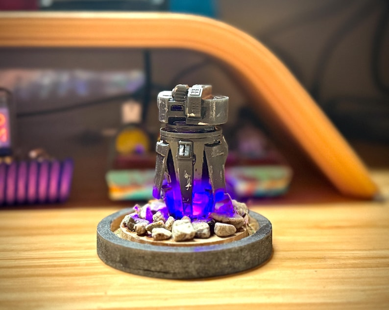 May include: A detailed, gray and black miniature sculpture with a futuristic design. The sculpture sits on a circular base with small rocks and emits a bright blue light. The base is made of a gray, textured material.