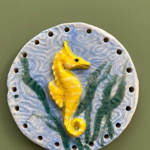 May include: A yellow ceramic seahorse with a blue and green background. The seahorse is in a standing position with its tail curled. The background has a swirling pattern.