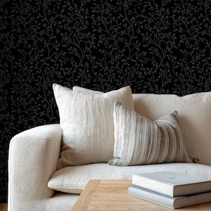 May include: A modern living room scene featuring a cream-colored sofa with several pillows, a wooden coffee table, and a decorative vase. The background is a black wallpaper with a subtle, repeating leaf pattern, creating a serene and stylish ambiance.