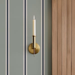 May include: A brass wall sconce with a lit white candle. The sconce has a circular base and a cylindrical candle holder. The background features vertical stripes in light blue, white, and navy. The right side shows wooden panels.