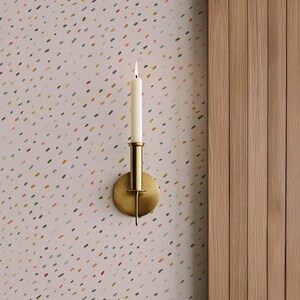May include: A brass wall sconce with a lit white candle. The sconce has a round base and a cylindrical candle holder. The background is a light pink wallpaper with colorful dotted patterns. To the right is a wooden panel.