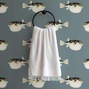 May include: A white hand towel with fringe hangs from a black metal ring. The towel is set against a teal background with a repeating pattern of pufferfish. The fish are white with brown spots and yellow eyes.