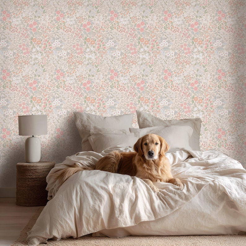This image showcases the wallpaper pattern in a bedroom setting. The pattern features a ditsy floral blossom design. The flowers are illustrated in shades of peach, coral, pink, and white against a beige background.