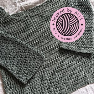 Può includere: Un top di uncinetto verde chiaro con un motivo a uncinetto. Il top ha maniche lunghe e una scollatura ampia. Un cerchio rosa con il testo "Hooked by Alli. It's a crochet pattern!" si trova nell'angolo in alto a destra dell'immagine.