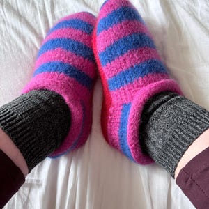 Può includere: Un paio di pantofole a righe rosa e blu. Le pantofole sono realizzate in un materiale morbido e soffice e poggiano su una superficie bianca. I calzini sono grigi.